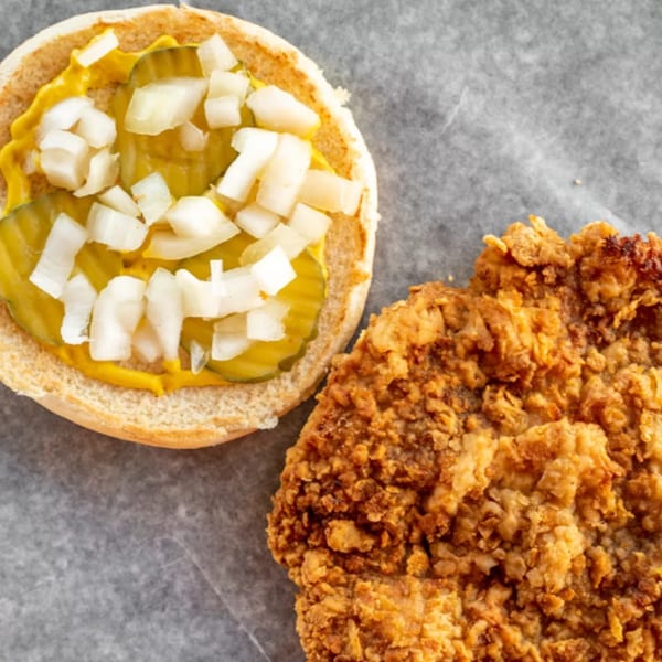 Top-down view of a crispy fried chicken patty and a burger bun with mustard, pickles, and onions.