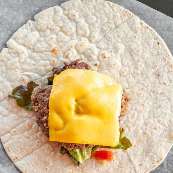 A burger patty with melted cheese, lettuce, and tomato on a tortilla, ready to be wrapped.